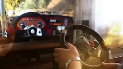 Eine Person hält ein Lenkrad vor einem Bildschirm, auf dem ein Armaturenbrett und eine Landschaft zu sehen sind.