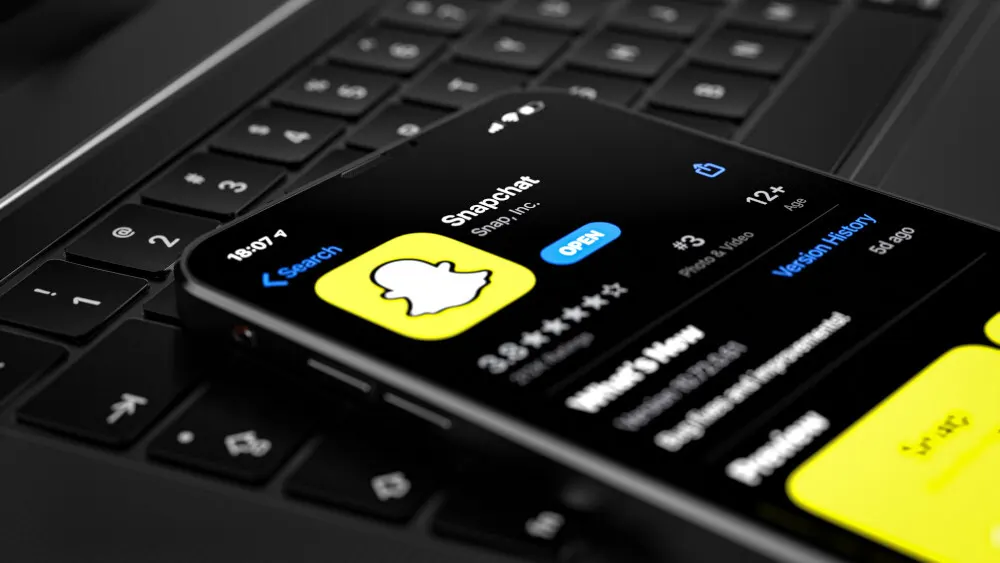 Handy liegt auf einer schwarzen Laptop-Tastatur. Auf dem Display ist das Snapchat-Symbol in Gelb und Weiß zu sehen.