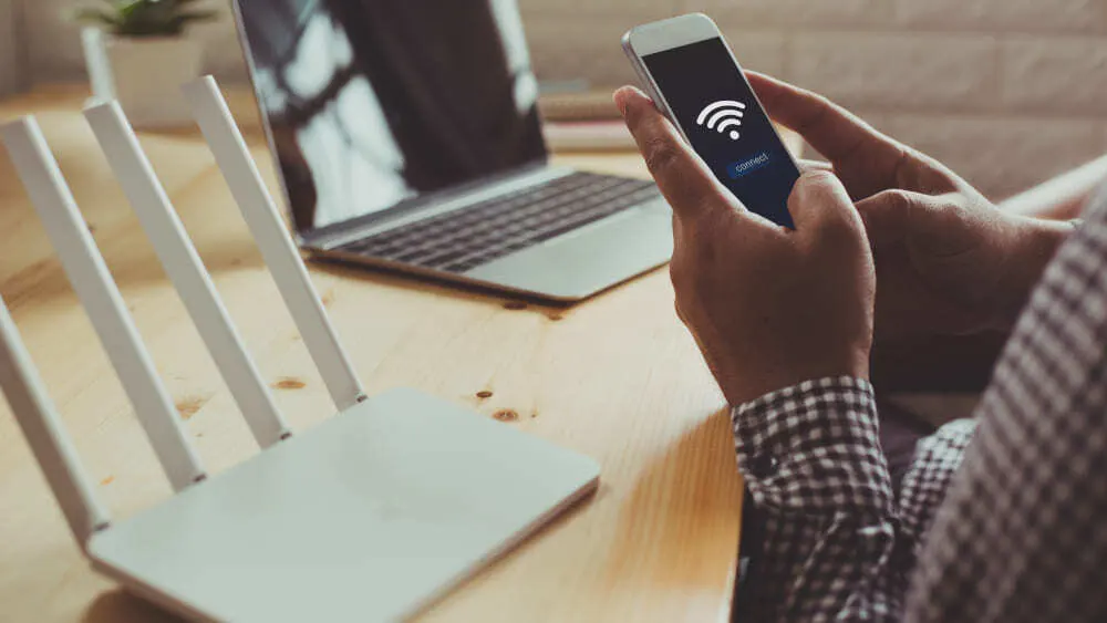 Auf einem Tisch liegen ein Laptop, ein WLAN-Router und ein Mobiltelefon in der Hand einer Person. Das Telefonsignal wird angezeigt.