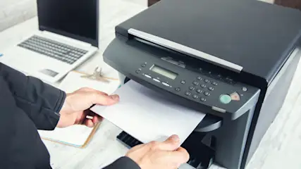 Eine Person legt Papier in einen Drucker. Im Hintergrund sind ein Laptop und ein Klemmbrett zu sehen.