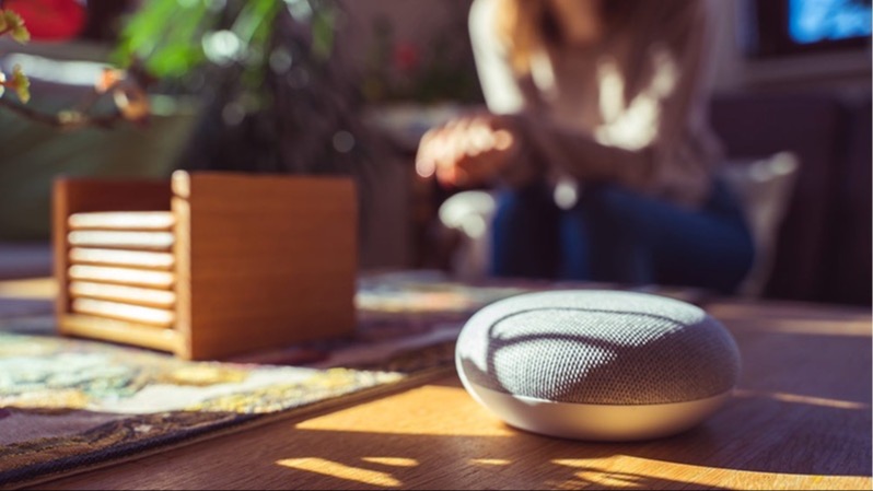 Ein grauer, runder Smart-Lautsprecher steht auf einem Tisch neben einem Holzuntersetzer. Im Hintergrund sitzt eine Person.