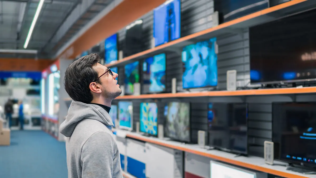 Mann mit Brille und grauem Kapuzenpullover betrachtet eine Wand voller eingeschalteter Fernseher im Geschäft.