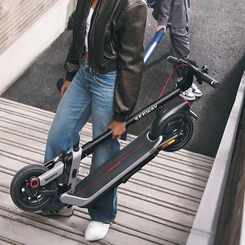 Eine Person trägt den zusammengeklappten SEGWAY Ninebot E3 Pro D E-Scooter eine Rampe hinauf, modernes schwarzes Design mit roten Akzenten.