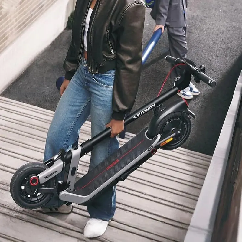 Eine Person trägt den zusammengeklappten SEGWAY Ninebot E3 Pro D E-Scooter eine Rampe hinauf, modernes schwarzes Design mit roten Akzenten.