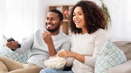 Ein Mann hält eine Fernbedienung, während eine Frau Popcorn isst. Beide lächeln und sitzen auf einem Sofa.