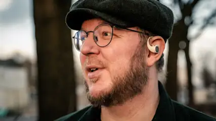 Ein Mann mit Bart und Brille trägt eine Schirmmütze und ein Hörgerät vor Bäumen.