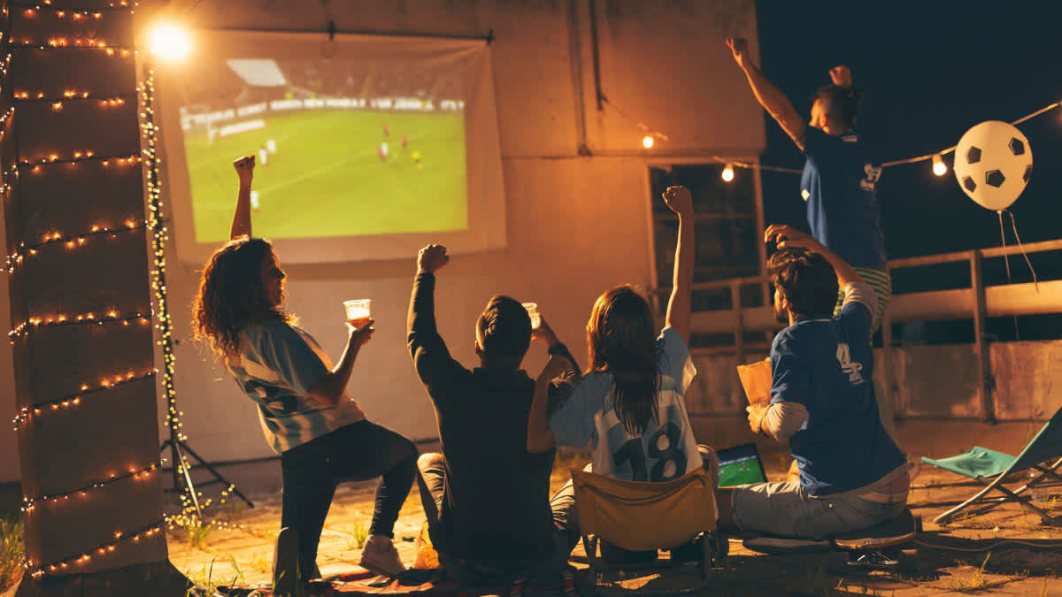 Mehrere Menschen jubeln gemeinsam während sie sich über eine Leinwand ein Fußballspiel anschauen.