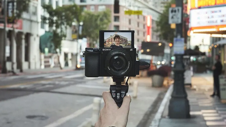 Eine Sony-Kamera auf einem Stativ wird von einer Hand gehalten, mit einer Person auf dem Kamerabildschirm vor einer Stadtstraße.