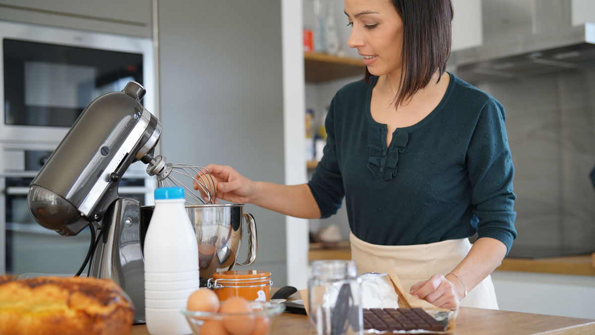Eine Frau schlägt ein Ei in die Schüssel einer Küchenmaschine, auf der Arbeitsplatte stehen weitere Zutaten. (Adobe Stock, Datei Nr.: 198071977)