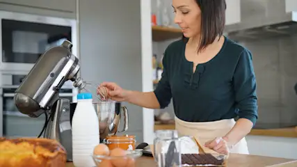Eine Frau schlägt ein Ei in die Schüssel einer Küchenmaschine, auf der Arbeitsplatte stehen weitere Zutaten. (Adobe Stock, Datei Nr.: 198071977)