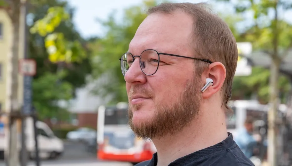 Ein Mann mit Bart und Brille trägt einen weißen Ohrhörer. Im Hintergrund sind Bäume und ein Bus zu sehen.