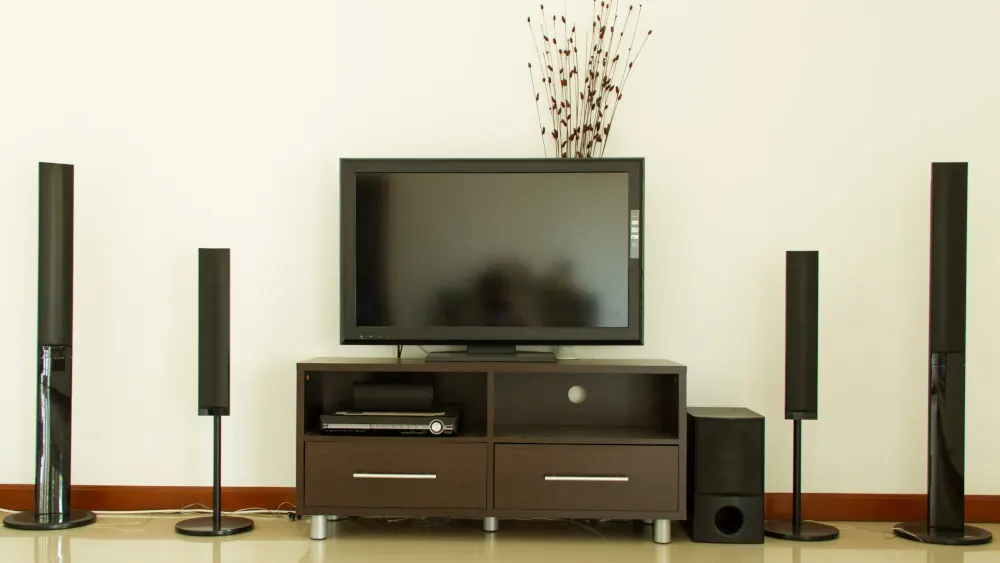 Ein Fernseher steht auf einem braunen Schrank mit Lautsprechern an den Seiten, vor einer weißen Wand.