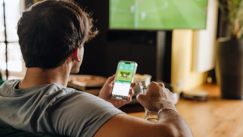 Ein Mann sitzt mit einem Smartphone vor einem Fernseher und schaut Fußball.