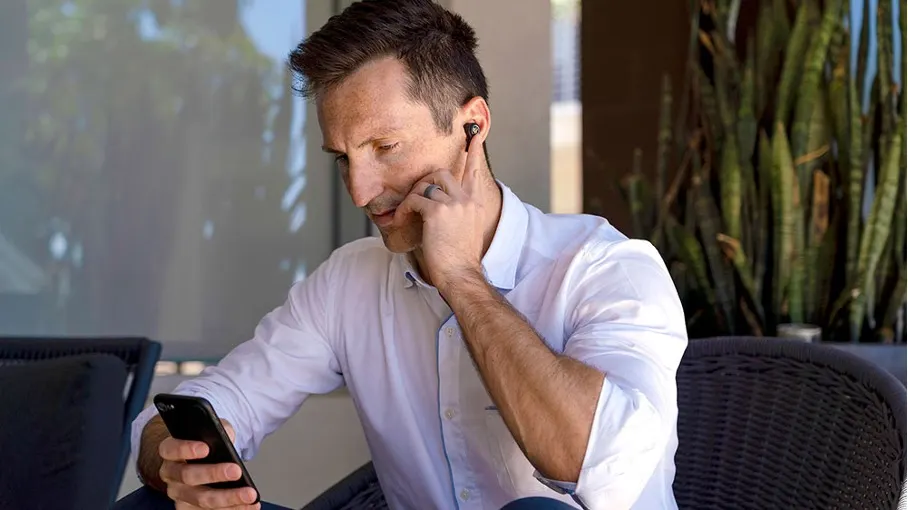 Ein Mann mit weißem Hemd sitzt und hält ein schwarzes Smartphone. Er berührt einen Ohrhörer. Im Hintergrund sind Pflanzen.
