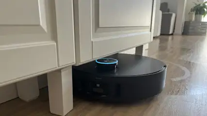 Ein schwarzer Saugroboter mit blauem Licht reinigt unter einem weißen Schrank auf einem Holzboden in einem Zimmer.