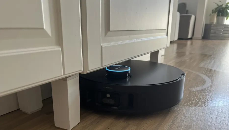 Ein schwarzer Saugroboter mit blauem Licht reinigt unter einem weißen Schrank auf einem Holzboden in einem Zimmer.