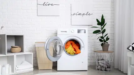 Waschmaschine mit offener Tür, gefüllt mit gelber und oranger Kleidung. Im Hintergrund sind Dekorationen und ein Regal zu sehen.