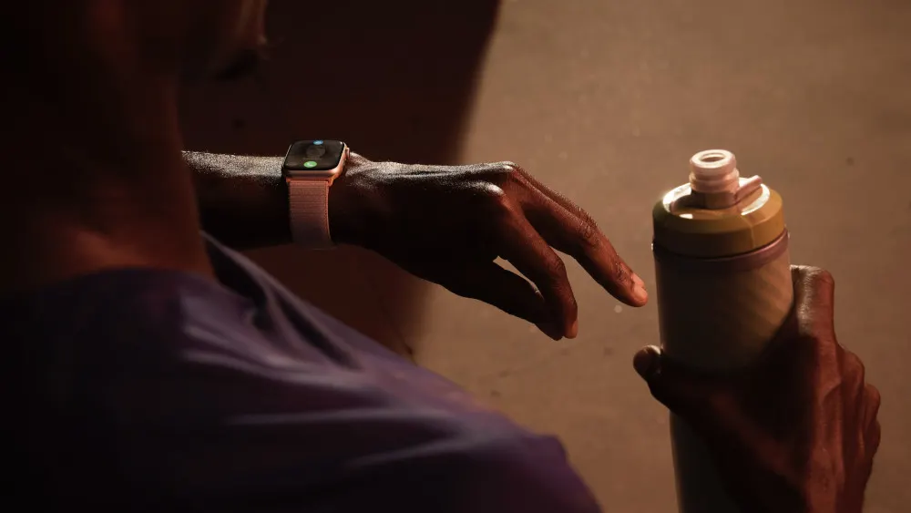 Eine Person mit einer Smartwatch berührt eine Wasserflasche. Die Flasche ist gelb, weiß und grau. Die Person trägt ein lila Hemd.