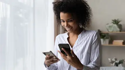Eine Frau im gestreiften Hemd hält zwei Smartphones vor einem Fenster mit Regalen und Pflanzen im Hintergrund.