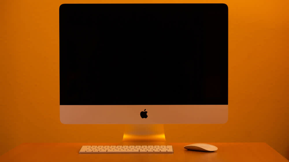 Ein iMac steht inklusive Tastatur und Maus auf einem Schreibtisch vor einem orangen Hintergrund. 