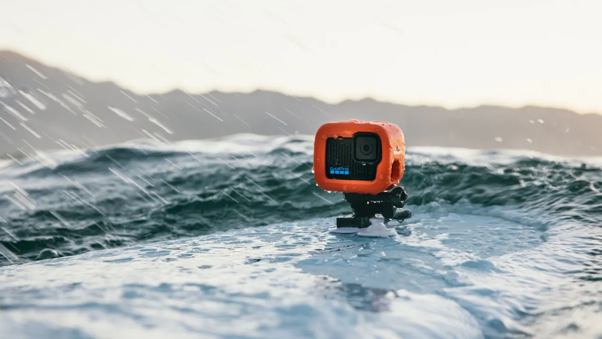 Eine orangefarbene GoPro-Kamera ist auf einem Surfbrett im Meer befestigt. Im Hintergrund sind Berge und Regentropfen sichtbar.