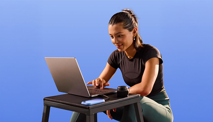 Person tippt am Laptop an kleinem Tisch; daneben Smartphone und schwarzes Zubehör, blauer Hintergrund.