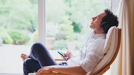 Ein Mann sitzt mit Kopfhörern in einem Stuhl und hält ein Telefon in der Hand. Draußen ist ein Fenster mit Grün.
