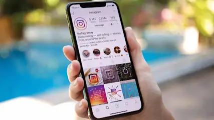 Ein schwarzes Smartphone zeigt die Instagram-App in der Hand. Das Display zeigt Beiträge, Symbole und Text vor einem verschwommenen Hintergrund.