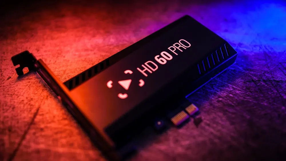Eine schwarze Elgato HD60 Pro Capture-Karte liegt auf einem strukturierten Untergrund, beleuchtet mit rotem und blauem Licht.