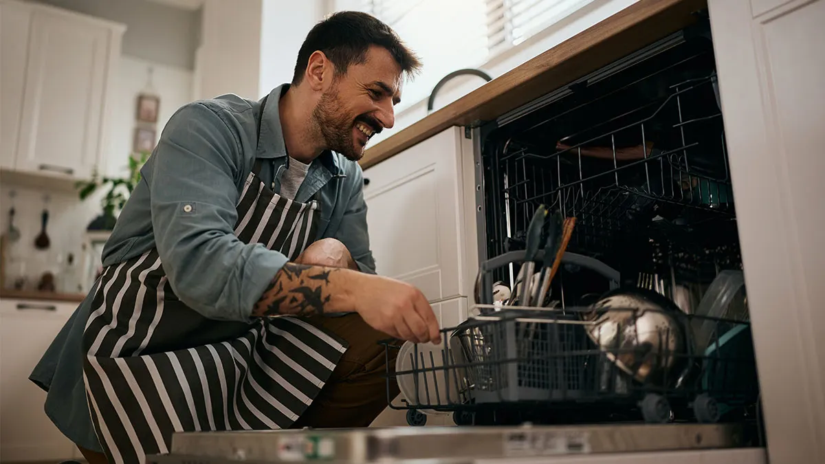 Ein lächelnder Mann mit gestreifter Schürze räumt Geschirr in eine Spülmaschine ein.