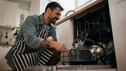 Ein lächelnder Mann mit gestreifter Schürze räumt Geschirr in eine Spülmaschine ein.