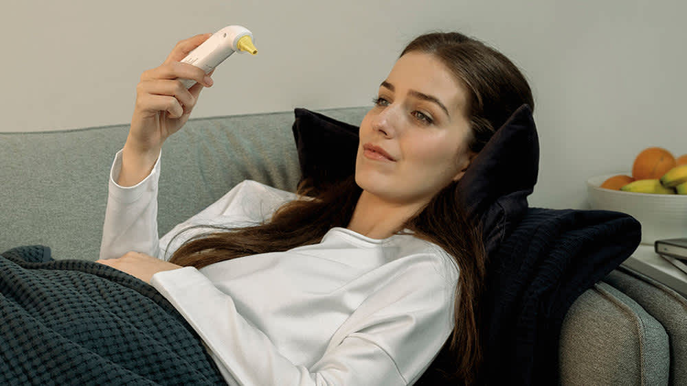 Eine kranke Frau liegt auf dem Sofa und liest ihre Körpertemperatur auf dem Braun ThermoScan 3 Fieberthermometer ab.