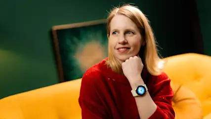 Eine Frau mit rotem Pullover sitzt auf einem gelben Sofa, eine Uhr am Handgelenk, vor einem grünen Hintergrund mit einem Gemälde.