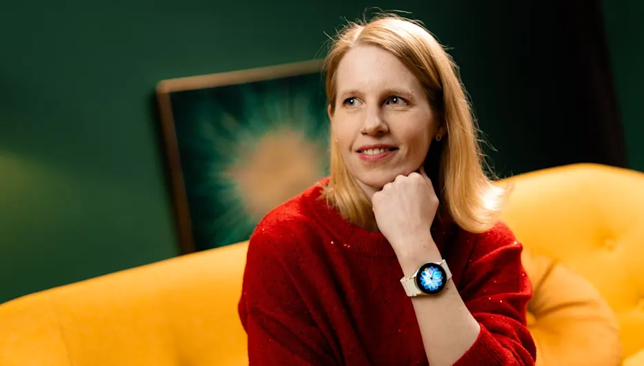 Eine Frau mit rotem Pullover sitzt auf einem gelben Sofa, eine Uhr am Handgelenk, vor einem grünen Hintergrund mit einem Gemälde.