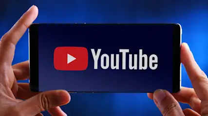 Hände halten ein schwarzes Smartphone mit dem YouTube-Logo auf blauem Hintergrund. Das Logo ist rot mit weißer Schrift.