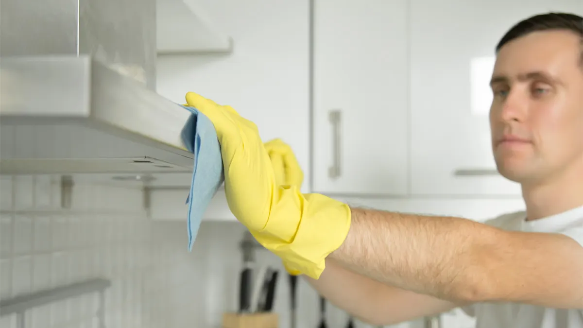 Mann putzt Dunstabzugshaube mit gelben Handschuhen und blauem Tuch. Weiße Schränke im Hintergrund.