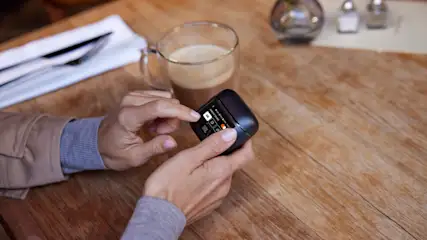 Auf einem Holztisch hält eine Hand ein JBL-Gerät. Daneben stehen ein Kaffee, Besteck und Salz- und Pfefferstreuer.