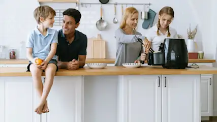In der Küche sitzt ein Kind mit einer Orange auf der Theke, daneben steht ein Mann. Eine Frau hantiert mit einer Fritteuse.