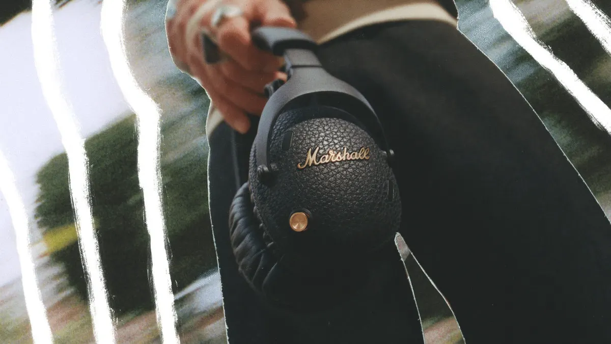 Eine Person hält schwarze Marshall-Kopfhörer mit goldenem Logo und Knopf vor einem verschwommenen Hintergrund.