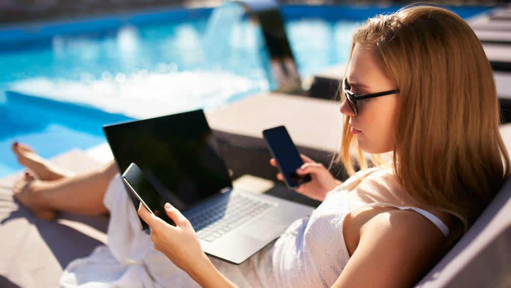 Eine Frau liegt am Pool mit zwei Smartphones und einem Laptop.