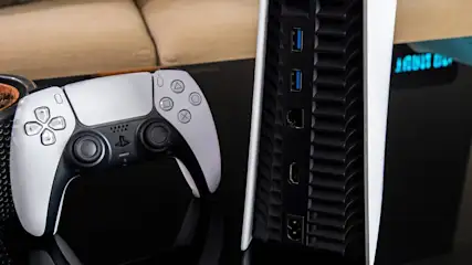 Ein grauer und schwarzer Playstation 5 Controller liegt neben einer Spielkonsole mit USB-Anschlüssen auf einem glänzenden, schwarzen Tisch.
