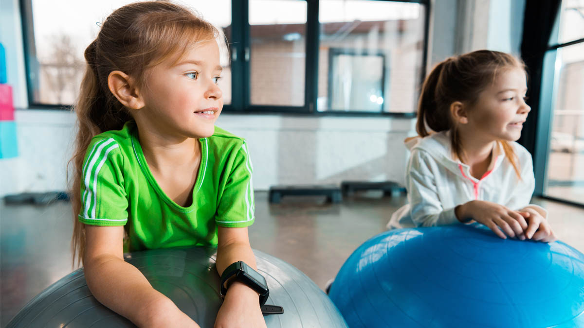 Zwei Mädchen in Sportkleidung lehnen auf Gymnastikbällen in einem Turnraum.
