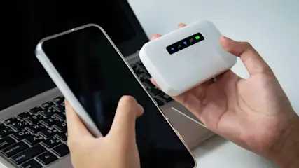 Eine Hand hält ein weißes Gerät mit grünen Lichtern, während die andere ein Mobiltelefon vor einem Laptop hält.