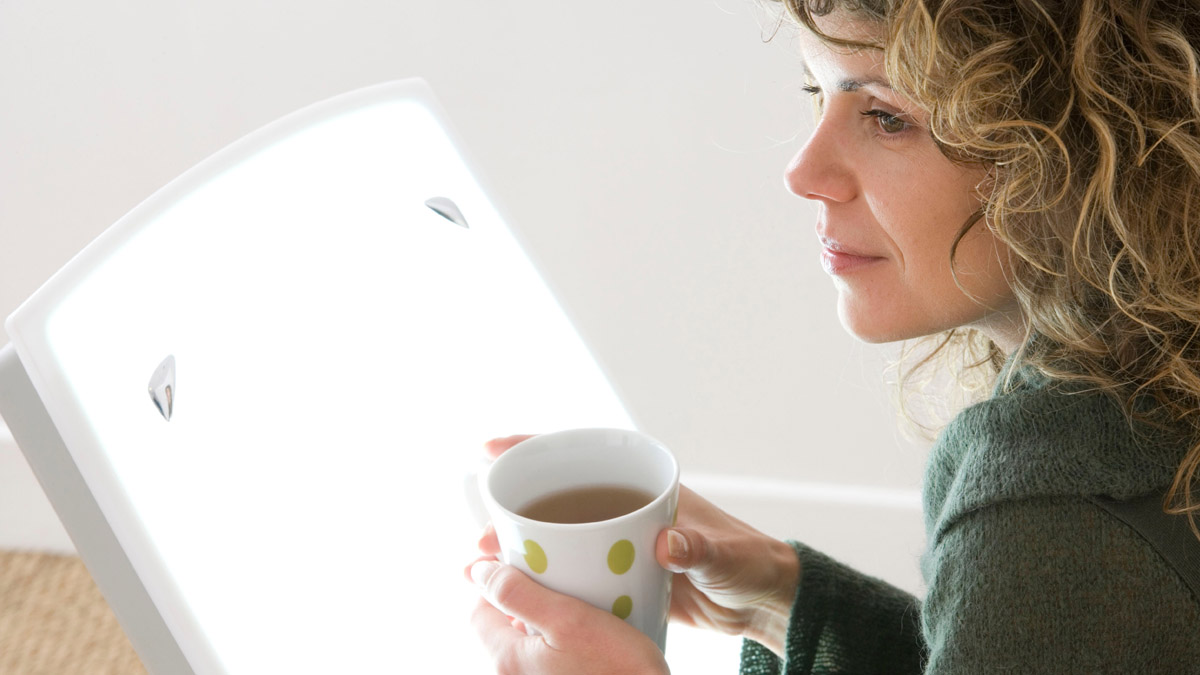 Eine Frau sitzt mit einer Kaffeetasse vor einer Tageslichtlampe.