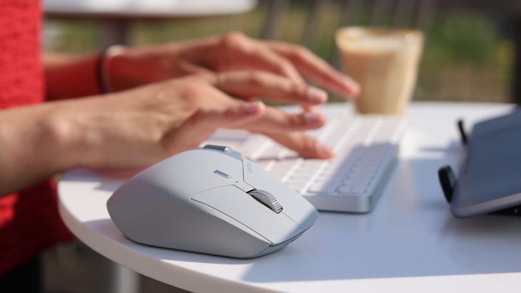 Nahaufnahme einer Hand auf einer weißen Tastatur neben einer hellgrauen Computermaus; im Hintergrund steht ein Glas Kaffee auf einem Tisch im Freien. Geben Sie Ihr Feedback zu BizChat ab.