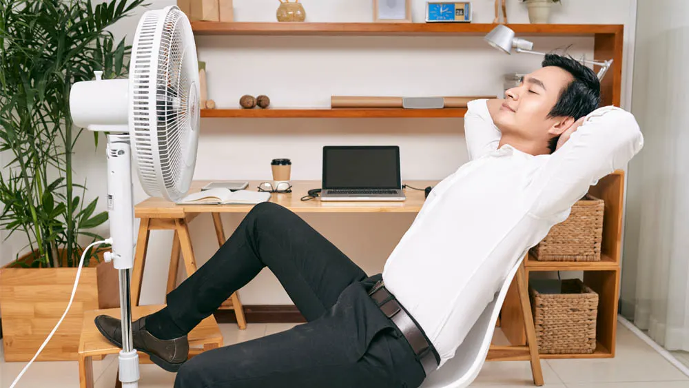 Ein Mann entspannt mit verschränkten Händen hinter dem Kopf vor einem Ventilator in einem Büro. Auf dem Tisch stehen ein Laptop und eine Kaffeetasse.