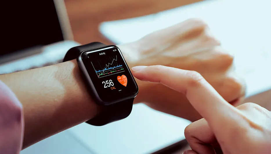 Eine Person tippt auf eine schwarze Smartwatch, die am Handgelenk getragen wird. Auf dem Bildschirm werden Kalorien und ein Herzsymbol angezeigt.