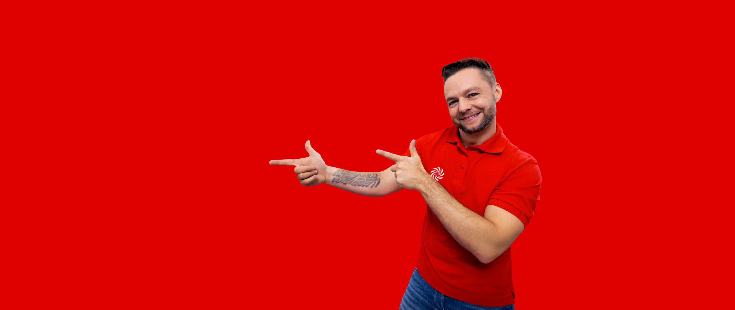 Ein Mann in rotem Poloshirt und Jeans zeigt auf rotem Hintergrund mit beiden Händen nach links. Ein Tattoo ist auf seinem Arm sichtbar.