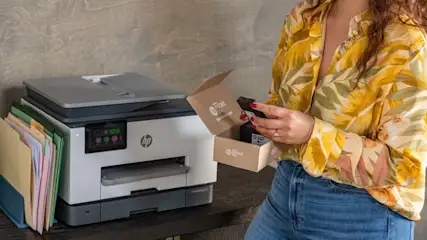Eine Person hält eine Tintenpatrone vor einem Drucker und einer Box mit dem Aufdruck "Instant Ink". Links davon stehen Ordner.