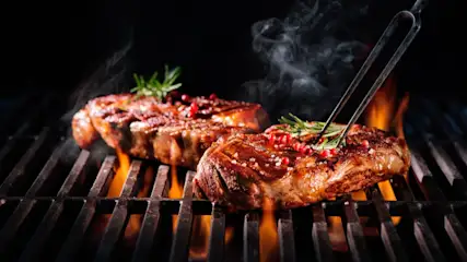 Zwei Steaks auf einem Grill mit Flammen. Ein Steak wird mit einer Gabel gehalten, garniert mit Rosmarin und roten Pfefferkörnern.